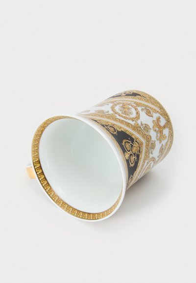 Taza de porcelana con patrones ornamentales en blanco y negro, detalles en dorado y borde dorado; mostrada boca abajo, revelando su interior blanco.