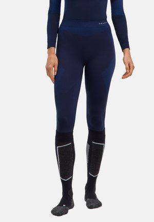 Person iført mørkeblå tætsiddende leggings og høje sorte og grå sportssokker, stående med hænderne afslappet langs siden.