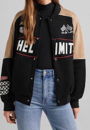 Frau trägt eine schwarz-beige Jacke mit Rennaufnähern, darunter karierte Flaggen und die Schriftzüge „HELL“ und „IMIT“ vorne, kombiniert mit hellen Jeans.