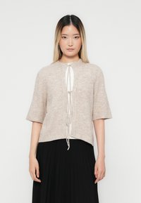 Frau mit langen, glatten Haaren, die eine beige Strickjacke mit kurzen Ärmeln trägt, die vorne über einem weißen Shirt und einem schwarzen plissee Rock gebunden ist.