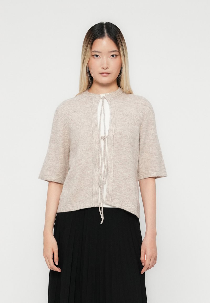 Frau mit langen, glatten Haaren, die eine beige Strickjacke mit kurzen Ärmeln trägt, die vorne über einem weißen Shirt und einem schwarzen plissee Rock gebunden ist.