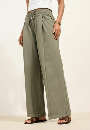 Kvinde iført olivengrønne bukser med vidde i benene, elastisk talje og snører, kombineret med brune flade sandaler og hvid top.