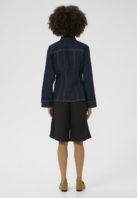 Džínová bunda v tmavě modré barvě s jasným prošíváním, přiléhavý střih, dlouhé rukávy a špičatý límec, v kombinaci s černými šortkami.