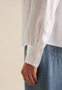 Chemise en lin blanche avec un poignet structuré, dotée de trois boutons. Associée à un denim bleu clair, offrant une coupe décontractée et une texture douce.