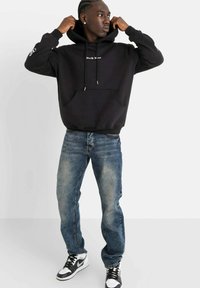 Felpa nera con tasca frontale e cappuccio regolabile, abbinata a jeans blu e scarpe da ginnastica nero e bianche. Il modello sta in piedi con le mani sul cappuccio.