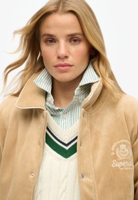 Karamelkleurige corduroy jas met een hoge kraag, denim-achtige knopen, gedragen over een crèmekleurige kabelgebreide trui met groene en marineblauwe strepen.