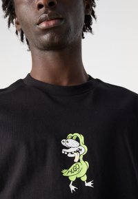 Camiseta de algodón negra con un vibrante personaje de dibujos animados de color verde, grandes ojos y una expresión juguetona, con el nombre "HUGO" estampado.