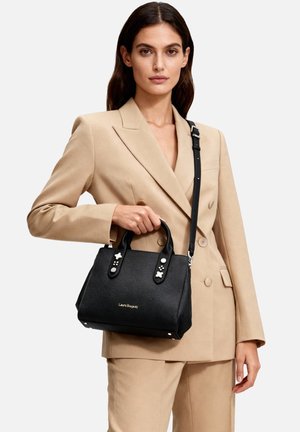 Donna in abito beige che tiene una borsa nera Laura Biagiotti con borchie, in piedi davanti a uno sfondo bianco neutro.