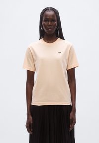 Lyse ferskenfarvet kortærmet t-shirt i bomuld med rund halsudskæring og lille grønt logo på brystet. Glat tekstur og afslappet pasform.