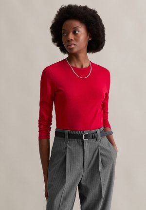 Top vermelho de mangas longas combinado com calças cinza risca de giz. A roupa inclui um cinto preto e um colar de prata, apresentando uma textura suave.