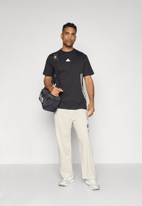 Homem de pé, vestindo uma t-shirt preta da Adidas, calças bege claras da Adidas, sapatilhas brancas, carregando uma mala de ombro preta da Adidas, contra um fundo simples.