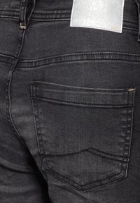 Jeans de mezclilla negra con una vista cercana del bolsillo trasero, que presenta costuras limpias y una etiqueta de cuero texturizada en la cintura.