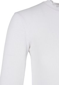 Detailní záběr na bílý žebrovaný dlouhý rukáv ukazující texturu látky a šití na rameni, paži a vysokém límci.