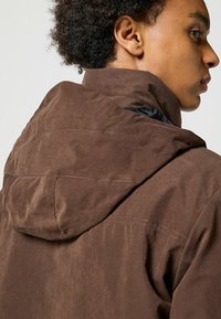 Giacca marrone, con texture e cappuccio, caratterizzata da dettagli cuciti e un rivestimento interno nero. Il design include un colletto alto e una forma affusolata.