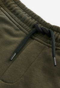 Pantalons de survêtement vert olive en tissu texturé, mettant en avant une taille froncée et un cordon noir avec embouts en plastique pour l'ajustement.