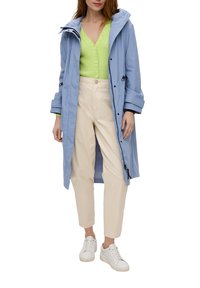 Lichtblauwe waterdichte jas met capuchon, vooraan met ritssluiting en koorden, gedragen over een groene geribbelde trui en beige broek. Witte sneakers.