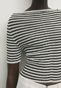 Mörkhyad person iklädd en svart- och vitrandig kortärmad topp och ljusa byxor mot en enfärgad bakgrund.