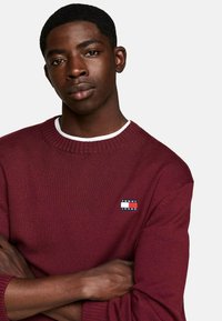 Suéter de punto burdeos con cuello redondo, que presenta un pequeño parche rectangular con el logo "TOMMY JEANS" en el lado izquierdo.