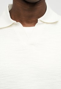 Εγγύς πλάνο λαιμού και άνω μέρος στήθους με σκούρο τόνο δέρματος, που φοράει λευκό πικέ πόλο πουκάμισο με γιακά και V-σχήμα στο λαιμό.