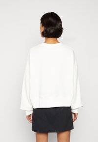 Vit oversized sweatshirt med sänkt axel, ribbade ärmslut och kantdetaljer. Bärs över en mörk kjol och visar en avslappnad silhuett.