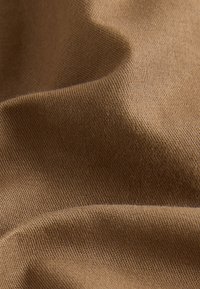 Tessuto marrone con trama intrecciata visibile e morbide pieghe che creano delicati giochi di ombre e luci.