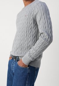 Grauer Cable-Knit-Pullover mit gerippten Bündchen und Saum. Rundhalsdesign, taillierte Passform, getragen über blauen Jeans. Unauffälliges Logo-Detail auf der Brust.