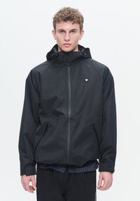 Jachetă waterproof neagră cu glugă ajustată, fermoar în față și două buzunare laterale. Dispune de un mic logo pe piept și manșete elastice.