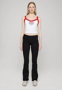 Witte cropped tanktop met rode bies en tekst, gecombineerd met zwarte uitlopende jeans. Model draagt grijs-witte sneakers. Neutrale achtergrond.