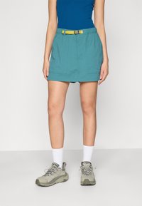 Calções turquoise com um cinto amarelo, apresentando dois bolsos frontais e um corte relaxado. Usados com sapatos cinzentos e meias brancas.