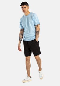Camiseta de algodón azul claro de manga corta, acompañada de pantalones cortos negros. El modelo lleva zapatillas blancas, mostrando un atuendo casual.