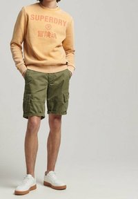 Světle oranžová mikina s nápisem "SUPERDRY", doplněná olivově zelenými cargo kraťasy. Bílé tenisky s hnědými podrážkami. Ležérní střih a design.