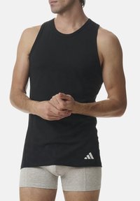 Černý bezrukávový top vyrobený z bavlny, s kulatým výstřihem a bílým logem Adidas v pravém dolním rohu. Viditelné šedé boxerky.