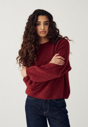 Giovane donna con capelli lunghi e ricci, che indossa un maglione rosso e jeans scuri, in piedi con le braccia incrociate davanti a uno sfondo neutro.