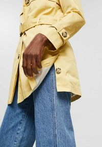 Trench-coat jaune avec une taille ceinturée, des poignets à boutons et des poches latérales ; associé à un jean bleu large.