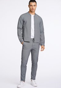 Man som bär en ljusgrå zip-up jacka, matchande smala byxor, vit t-shirt och vita sneakers, stående mot en enkel vit bakgrund.