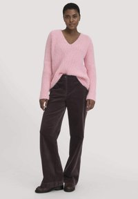 Rosa gerippter V-Ausschnitt-Pullover mit langen Ärmeln, kombiniert mit dunklen Cordhosen in weiter Beinform. Die Schuhe sind braun mit einer flachen Sohle.