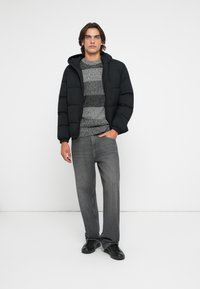 Giovane uomo che indossa una giacca imbottita nera, un maglione a righe grigio, jeans grigi e sneakers nere, in piedi con una mano in tasca.