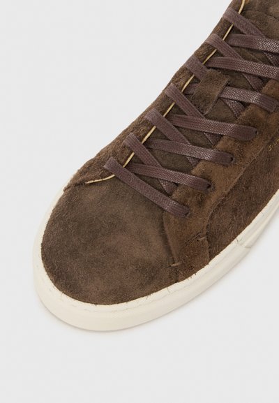 Brown suede sneaker featuring a rounded toe, textured upper, and flat brown laces, complemented by a cream-colored rubber sole.