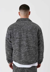 Uomo con capelli corti che indossa una giacca a scacchi bianca e nera e pantaloni abbinati, in piedi di spalle su uno sfondo uniforme.