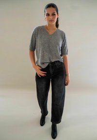 Femme aux cheveux bruns en queue de cheval portant un pull gris à manches courtes, un jean noir taille haute, des bottes noires et des bijoux en or sur un fond uni.