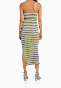 Femme portant une robe sans manches, arrivant aux genoux, à rayures vertes, bleues et jaunes avec une fente sur le côté, associée à des sandales à talons hauts bleu clair.