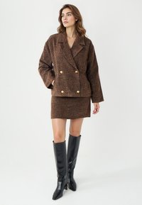 Brauner strukturierter, doppelreihiger Blazer mit goldenen Knöpfen, kombiniert mit einem passenden Rock und ergänzt durch kniehohe schwarze Lederstiefel.