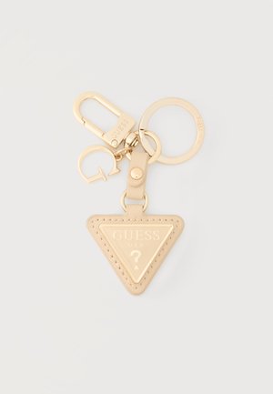 Μπεζ και χρυσό μπρελόκ Guess με ετικέτα λογότυπου σε σχήμα τριγώνου, επώνυμο κρίκο κλειδιών, κλιπ και μικρό γούρι με το γράμμα "G" σε φωτεινό φόντο.