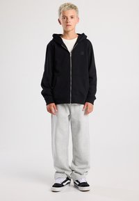 Schwarze Zip-Hoodie mit einer Fronttasche, kombiniert mit hellgrauen Jogginghosen und schwarzen Turnschuhen mit weißen Akzenten. Das Modell steht aufrecht.