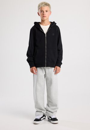 Sudadera negra con cremallera y bolsillo frontal, combinada con pantalones de chándal gris claro y zapatillas negras con acentos blancos. El modelo está de pie de manera recta.