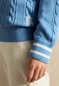 Pulover tricotat de culoare albastru deschis, cu manșete cu margini ondulate, cu dungi albe, logo brodat mic pe față și motive texturate.