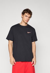 Jeune homme avec des cheveux bouclés portant un T-shirt noir Nike avec un logo orange et un short rouge, sur un fond gris clair uni.