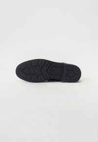 Suola di scarpa nera in gomma con un motivo a battistrada testurizzato, caratterizzato da scanalature e un design a forma di freccia per trazione e stabilità.
