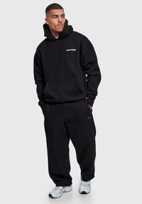 Sweat à capuche noir et joggeurs assortis en molleton, avec une poche avant et un logo en accent. Le modèle porte des baskets blanches.