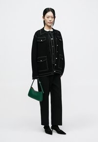 Black wool jacket with white stitching, two front pockets, black trousers, and a green handbag. Model wears pointed black shoes.